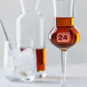 Grappaglas mit Gravur „24“ vor Karaffe und Glas mit Eiswürfeln