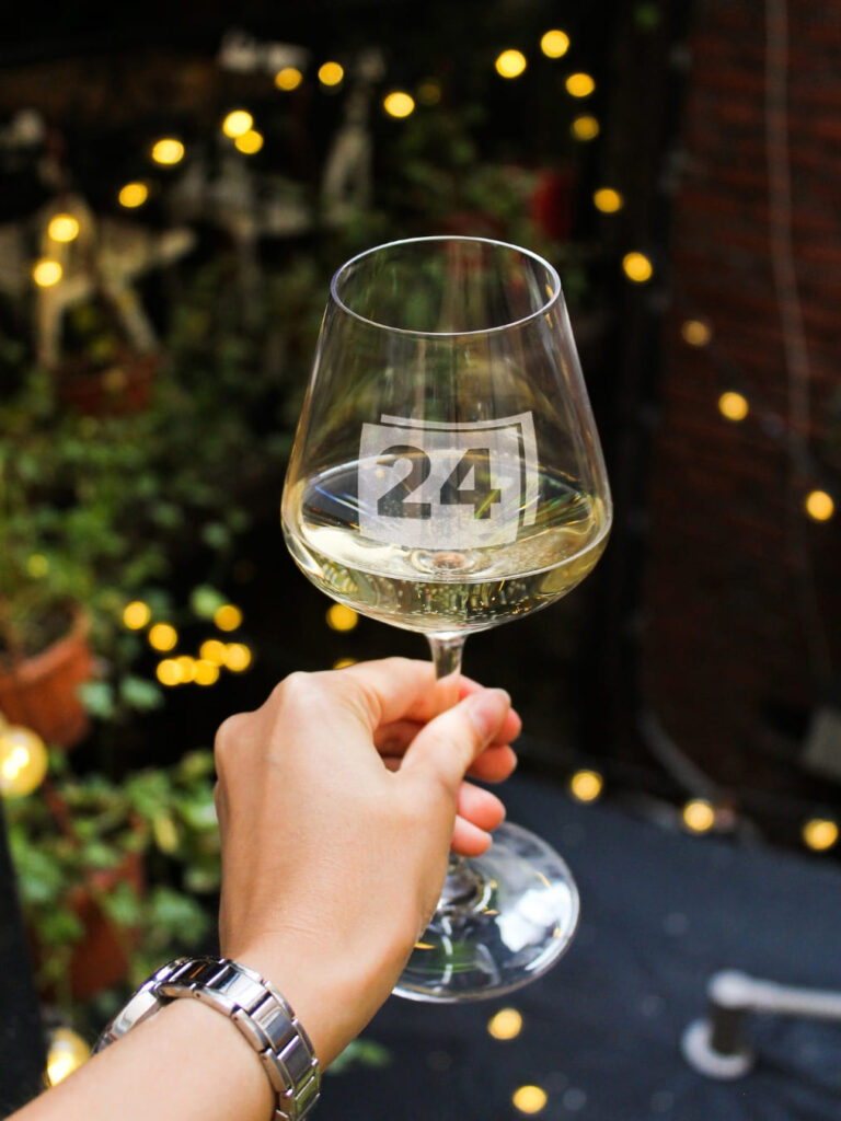 Weißweinglas mit Gravur „24“, in Hand gehalten mit Lichterkette