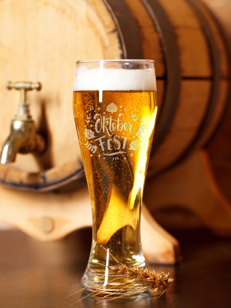 Weizenglas mit Gravur „Oktoberfest“, gefüllt mit Bier vor Holzfass