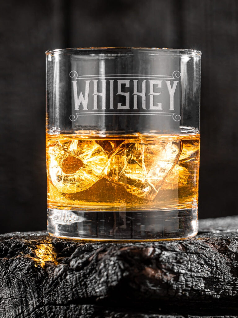 Whiskeyglas mit Gravur „Whiskey“ und Eiswürfeln auf dunklem Holz
